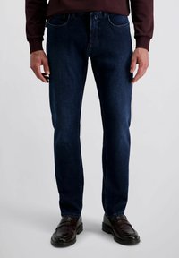 Man in een donkerblauwe slim-fit jeans, bruine leren loafers en een maroon longsleeve shirt, staand tegen een egale lichte achtergrond.