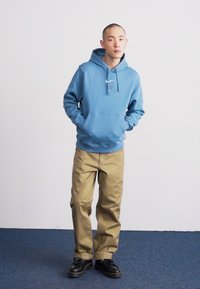 Sweatshirt azul-claro com capuz, com um bolso frontal e logótipo, combinado com calças caqui e sapatos pretos, de pé sobre um tapete azul contra uma parede branca.