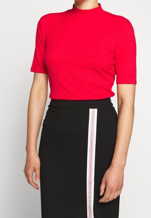 Femme portant un haut à manches courtes rouge à col roulé et une jupe noire avec une bande verticale blanche affichant un texte géométrique rouge.