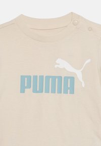 Μπεζ πουκάμισο Puma με μπλε κείμενο "PUMA" και λευκό λογότυπο πούμα που πηδά, με κουμπιά κούμπωμα στο δεξί ραφτό του ώμου.