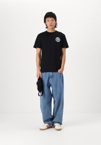 Black cotton t-shirt with a round logo on the chest, paired with loose blue denim jeans and white sneakers with red stripes.