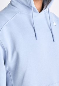 Sweat à capuche bleu clair avec cordons, manches raglan et petit logo blanc sur la poitrine, porté par une personne à la peau brune.