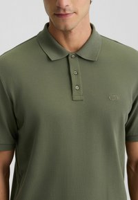 Olivgrünes Poloshirt aus strukturiertem Stoff. Verfügt über einen klassischen Kragen, eine Drei-Knopf-Leiste und ein dezentes Logo auf der linken Brust.