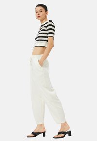 Szydełkowy crop top w czarno-białe paski zestawiony z białymi spodniami w wysokim stanie; model nosi czarne klapki. Gładki materiał i luźny krój.