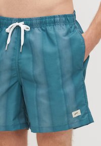 Tyrkysové plavecké šortky z ľahkého materiálu s elastickým pásom, bielou šnúrkou a bočnými vreckami; obsahujú malú nášivku s logom.