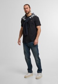 Schwarzes kurzärmliges Hemd, dunkelblauer Pullover um den Hals gebunden, blaue Jeans und cremefarbene Sneakers mit beigen Akzenten. Dezente TaschenDETAIL auf dem Hemd.