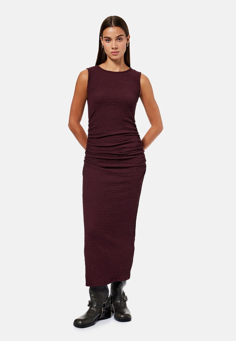 Femme debout, les mains dans le dos, vêtue d'une robe maxi sans manches texturée de couleur violet foncé et de bottines noires sur fond blanc.