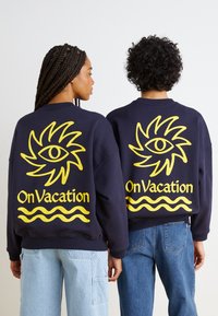 Τα ναυτικά oversized φούτερ διαθέτουν μια μεγάλη κίτρινη γραφική σχεδίαση ενός ματιού και το κείμενο "Σε Διακοπές" με κυματιστές γραμμές στο κάτω μέρος.