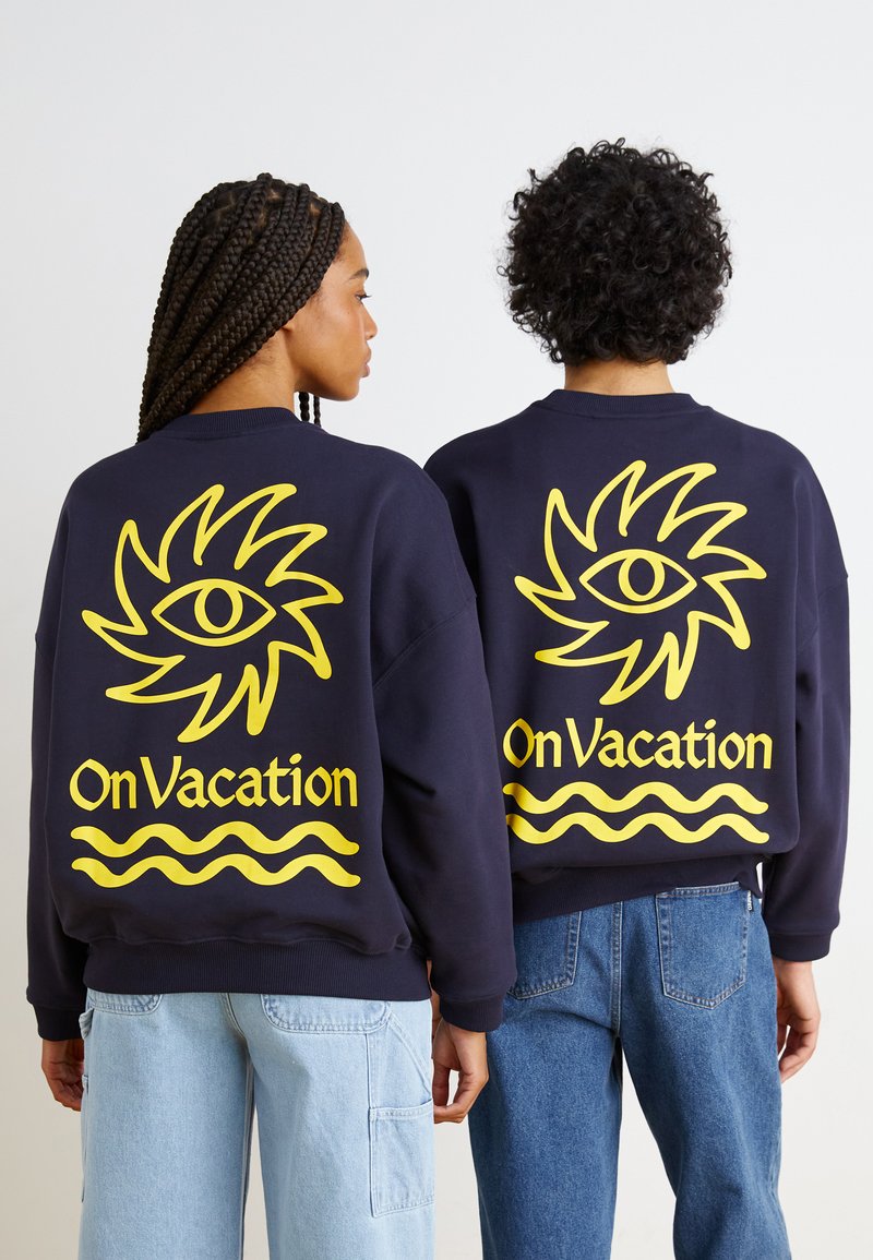 Τα ναυτικά oversized φούτερ διαθέτουν μια μεγάλη κίτρινη γραφική σχεδίαση ενός ματιού και το κείμενο "Σε Διακοπές" με κυματιστές γραμμές στο κάτω μέρος.