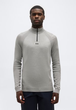 Mladý muž nosiaci svetlošedý pletený pulóver s dlhým rukávom, čiernym polozipsom a logom "BOSS" na hrudi, stojaci pred jednoduchým pozadím.