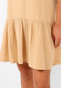 Robe beige clair en tissu texturé, avec un ourlet évasé court et des détails froncés à la taille. Le tissu semble doux et froissé.