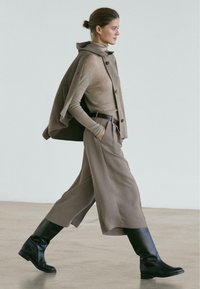 Beige langärmeliges Oberteil unter einem grauen Umhang, weite beige Hosen und kniehohe schwarze Lederstiefel mit flacher Sohle, gehend auf einem glatten Boden.