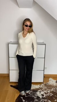 Cămașă lungă cu mânecă și decolteu în V, cu dungi crem, asortată cu pantaloni negri evazați și încălțăminte argintie. Accesoriile includ ochelari de soare negri și un lanț de aur.