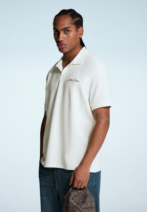 Polo in cotone avorio con una texture morbida. Presenta un colletto classico e un logo ricamato discreto sul petto. Abbinato a jeans blu.