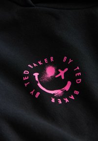 Musta kangas tekstuuripinnalla, jossa on vaaleanpunainen graafinen kuvio, mukaan lukien hymyilevä kasvot ja teksti "BY TED BAKER."