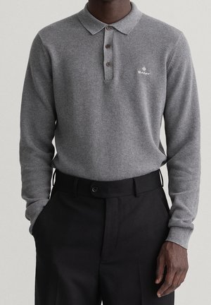 Homme portant un polo en maille gris à manches longues avec des boutons et un pantalon noir taille haute, mains dans les poches.
