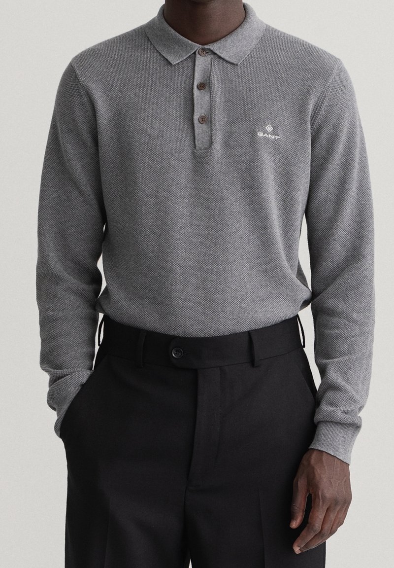 Homme portant un polo en maille gris à manches longues avec des boutons et un pantalon noir taille haute, mains dans les poches.