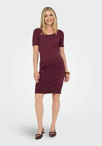 Femme souriante portant une robe ajustée bordeaux mi-longue à manches courtes et des talons assortis, posant devant un fond clair uni.