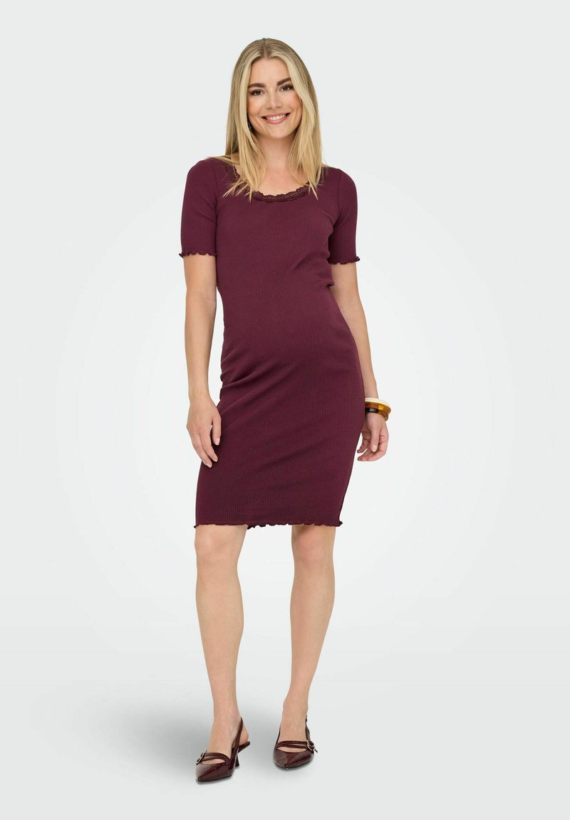 Femme souriante portant une robe ajustée bordeaux mi-longue à manches courtes et des talons assortis, posant devant un fond clair uni.