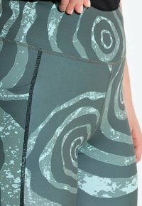 Leggings de cintura alta en verde oscuro con un patrón de espiral en azul claro. Tejido elástico con costuras cosidas. Longitud hasta la mitad del muslo, diseño ajustado.
