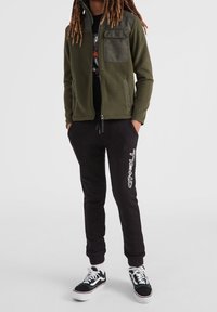 Oliven grøn fleecejakke med en kontrasterende tekstureret lomme, parret med sorte sweatpants med et hvidt logo og klassiske sorte sneakers.