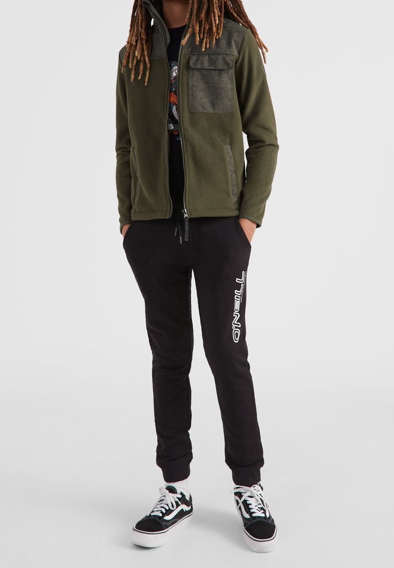 Oliven grøn fleecejakke med en kontrasterende tekstureret lomme, parret med sorte sweatpants med et hvidt logo og klassiske sorte sneakers.