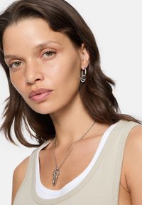 Smycken i silvermetall har en kedjeörhänge och ett hängande halsband med sammanlänkade ringar, tillsammans med en beige linne.