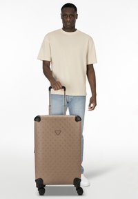 Hombre con camiseta beige y vaqueros sostiene el asa de una gran maleta marrón con un diseño en patrones y protectores de esquina negros, sobre un fondo blanco.