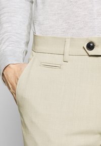 Hand i fickan på beige högmidjade byxor med en svart knapp och bälteslinga, kombinerat med en ljusgrå långärmad topp.