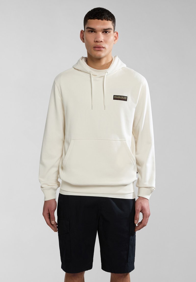 Sudadera crema con un bolsillo frontal, capucha con cordones y un pequeño parche de logo. Lleva puesta unos pantalones cortos cargo negros. Tela suave, diseño casual.