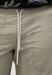 Χακί παντελόνι με κορδόνι και λευκό σχέδιο, με ορατή ραφή, φορεμένο κάτω από μαύρο τοπ με λουλούδια.