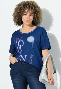 Žena s kudrnatými vlasy, oblečená v modrém tričku s potiskem a tmavých džínách, držící malou stříbrnou kabelku na bílém pozadí.