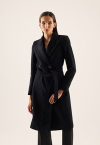 Femme aux cheveux raides et foncés portant un manteau en laine noir ceinturé avec de larges revers, debout devant un fond clair.