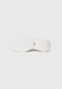 Suela de zapatilla deportiva blanca con un patrón de banda de rodadura texturizado y multidireccional, y un acento triangular rojo en el centro.