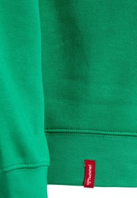 Grüner Stoff mit sichtbaren Nähten und einem roten Etikett mit der Aufschrift „hummel“, das am unteren Rand des Kleidungsstücks angenäht ist.