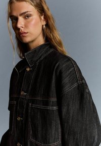 Schwarze Jeansjacke mit doppelten Brusttaschen, goldenen Knöpfen und kontrastierenden Nähdetails. Verfügt über einen hohen Kragen und eine lockere Passform.