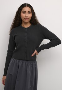 Cardigan cu dungi de culoare gri închis, cu guler rotund, închidere cu nasturi și mâneci lungi, asortat cu o fustă lejera, plisată, de culoare gri închis.