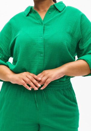 Žena oblečená v zelené košili na knoflíky s ohrnutými rukávy a zelených kalhotách, ruce jemně sepnuté v pase.