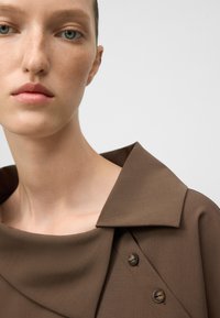 Blusa marrone con un ampio colletto e piccoli bottoni. Tessuto morbido con dettagli in drappeggio attorno al collo e alle spalle.