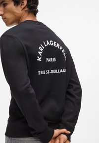 Sudadera negra con mangas largas, puños acanalados y cuello redondo. La parte trasera presenta el texto blanco "KARL LAGERFELD PARIS 21 RUE ST-GUILLAUME."