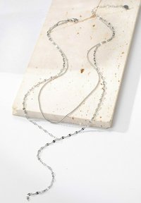 Silberne Kettenhalskette mit verschiedenen Gliedformen, die eine glatte Textur und einen glänzenden Finish aufweist, auf einer hellen Steingrundfläche liegend.
