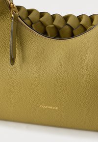 Oliivirohelise tekstuuriga nahast käekott punutud sangadega, kuldse tõmblukuga ja esiküljel kullas kuldse COCCINELLE logoga.