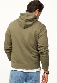 Olivgrüner Hoodie mit Kängurutasche, ausgestattet mit einer Kordelzugkapuze, gerippten Bündchen und Saum, getragen über einem weißen Shirt.