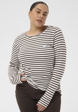 Frau mit dunklen Haaren in einem niedrigen Dutt, trägt ein weiß-braun gestreiftes Langarmshirt und dunkle Hose, posiert mit einer Hand in der Tasche.