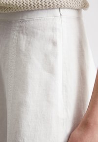 Weißer Leinenrock mit einer glatten Textur, dezenten Falten und einem seitlichen Nahtdetail. Kombiniert mit einem strukturierten hellbeigen Strickoberteil.