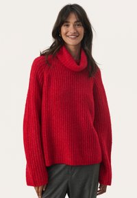 Rode gebreide trui met een robuuste textuur, coltrui en lange, losse mouwen. Draagt bij grijze broek, tegen een effen achtergrond.