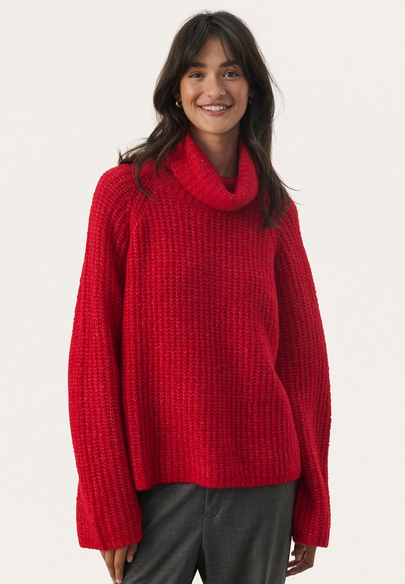 Rode gebreide trui met een robuuste textuur, coltrui en lange, losse mouwen. Draagt bij grijze broek, tegen een effen achtergrond.