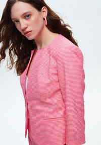 Blazer a scacchi rosa e bianco con un design aderente, spalle strutturate e un colletto piccolo; presenta una chiusura con un bottone singolo e tasche laterali.