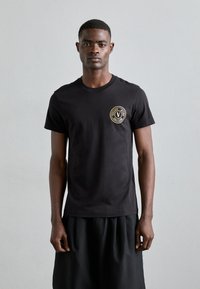 Μαύρη βαμβακερή μπλούζα με στρογγυλή λαιμόκοψη και κυκλικό χρυσό λογότυπο στην αριστερή πλευρά του στήθους. Φοριέται με μια μαύρη, φαρδιά φούστα.