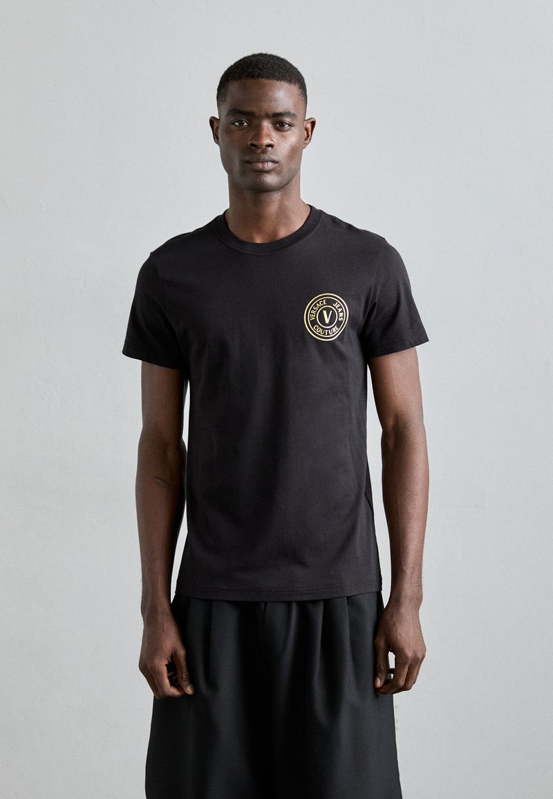 Μαύρη βαμβακερή μπλούζα με στρογγυλή λαιμόκοψη και κυκλικό χρυσό λογότυπο στην αριστερή πλευρά του στήθους. Φοριέται με μια μαύρη, φαρδιά φούστα.